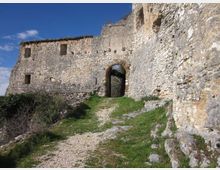 Mura in pietra di una fortificazione in rovina con un grande arco d’ingresso e piccole aperture, affacciate su un sentiero sterrato con tratti d’erba. La scena è all’aperto, su un pendio roccioso, sotto un cielo azzurro.