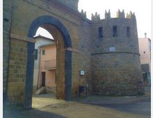 Antica porta urbana in pietra con grande arco d’ingresso e torre cilindrica merlata adiacente. Sullo sfondo si vedono edifici residenziali e una strada lastricata che attraversa il varco.