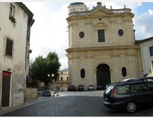 Facciata di una chiesa storica in pietra chiara su una piazza lastricata, con grande portale ad arco centrale, finestre rotonde e frontone classico. Ai lati si vedono edifici residenziali, alcune auto parcheggiate e alberi vicino al margine della piazza.