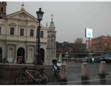 Piazza urbana con una chiesa storica dalla facciata classica e un’edicola monumentale con croce; in primo piano si vedono lampione, bicicletta e dissuasori. Sullo sfondo compaiono una strada con auto, un ponte e palazzi lungo il fiume.