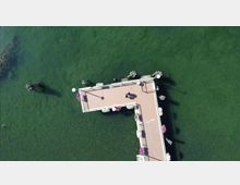 Vista dall’alto di un molo a forma di “L” che si estende su un lago verde, con ringhiere e fioriere lungo i bordi e alcune piccole strutture alle estremità. L’acqua circonda completamente la passerella, con ombre proiettate sul fondale.