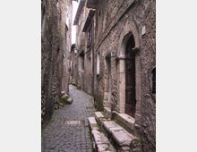 Stretto vicolo medievale in pietra con pavimentazione in ciottoli, fiancheggiato da alte mura e facciate in muratura grezza. Sul lato destro si vede un portale ad arco con porta in legno e gradini in pietra, con finestre e lampioni lungo il passaggio.