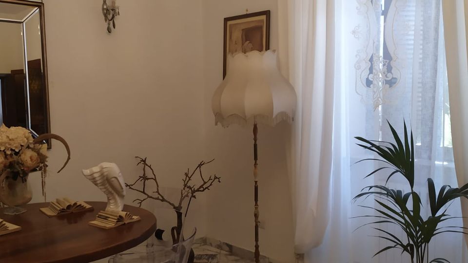 Interno di una sala da pranzo con pavimento in marmo, tavolo rotondo in legno e sedie trasparenti, illuminata da un lampadario di cristallo. Accanto a una grande finestra con tende bianche ci sono una pianta in vaso e una lampada da terra, con specchio e applique a parete.