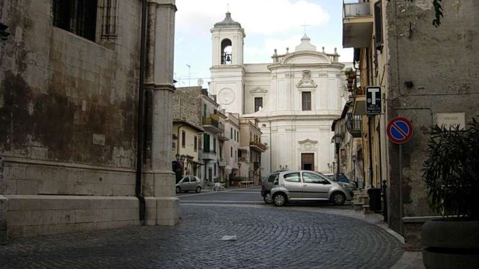 Strada acciottolata in un centro storico che conduce a una chiesa con facciata barocca chiara e campanile sullo sfondo. Ai lati si vedono edifici in pietra e intonaco, balconi e segnaletica stradale.