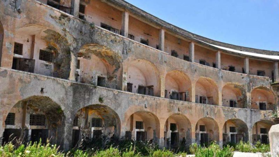 Edificio storico in rovina con facciata curva a più piani, caratterizzata da file di arcate e balconate con intonaco scrostato e finestre sbarrate. In primo piano si vede un cortile con vegetazione incolta e pavimentazione consumata sotto un cielo sereno.