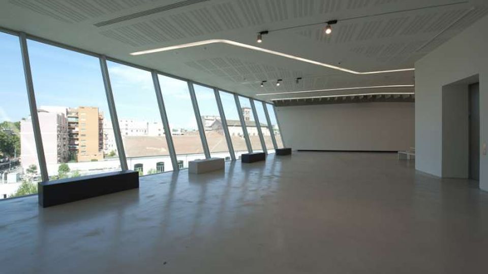 Ampia sala interna moderna e vuota, con pavimento chiaro e soffitto a pannelli con illuminazione lineare e faretti. Sul lato sinistro una lunga parete di grandi vetrate inclinate offre vista su edifici urbani e fa entrare molta luce naturale.