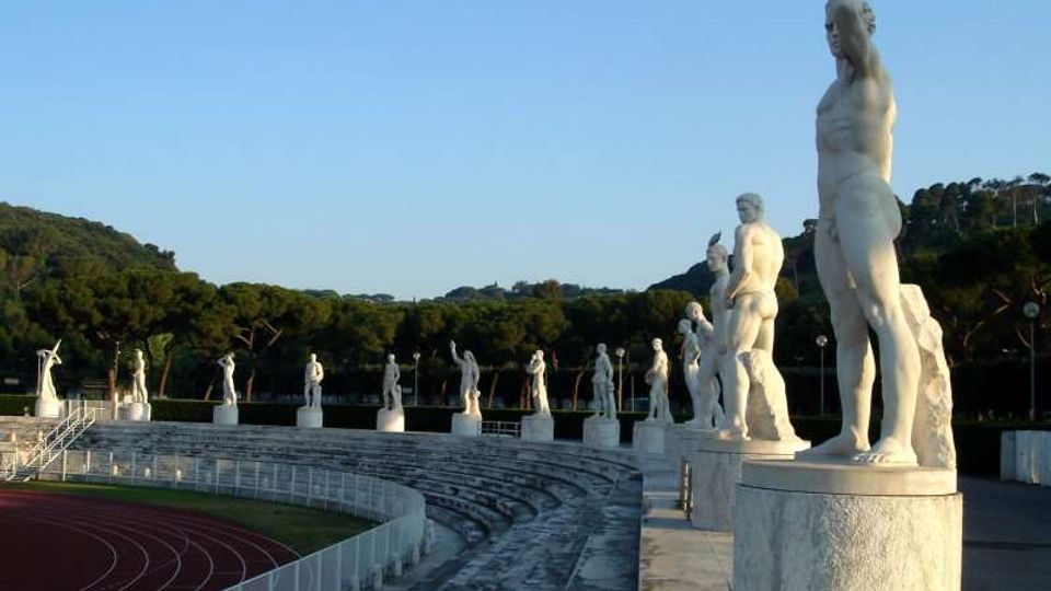 Anfiteatro all’aperto con gradinate in pietra che costeggiano una pista di atletica, affiancate da una fila di grandi statue bianche su piedistalli lungo il bordo superiore. Sullo sfondo si vedono colline alberate e cielo sereno.