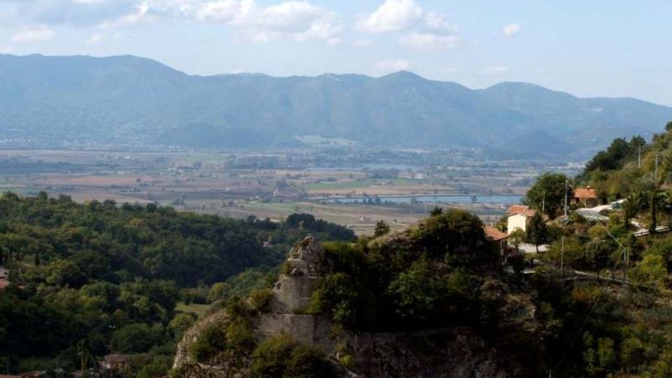 Veduta panoramica da una collina con vegetazione e rocce, con alcune case e una strada sul versante. Sullo sfondo si estende una pianura con campi e corsi d’acqua, chiusa da una catena montuosa sotto un cielo parzialmente nuvoloso.