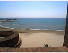 Veduta dall’alto di una spiaggia sabbiosa e del mare calmo, con frangiflutti e un molo che si estendono verso l’orizzonte. In primo piano si vede una struttura in muratura curva e un parapetto in mattoni.