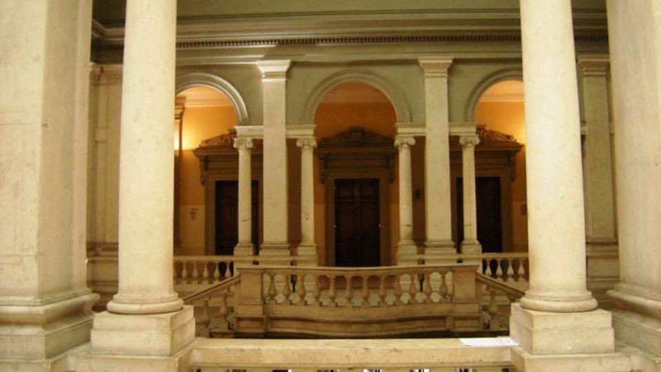 Interno di un cortile coperto con colonne in pietra e logge ad archi su due livelli, con balaustre e portali in legno sullo sfondo. Illuminazione calda sulle pareti e dettagli classici lungo i cornicioni.