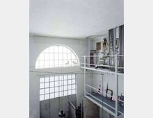 Interno di un loft a doppia altezza con soppalco e ringhiere bianche, grandi finestre a griglia e una finestra ad arco che illuminano lo spazio. Sul soppalco si vedono opere e cornici appoggiate e pareti con quadri, in un ambiente in stile industriale.