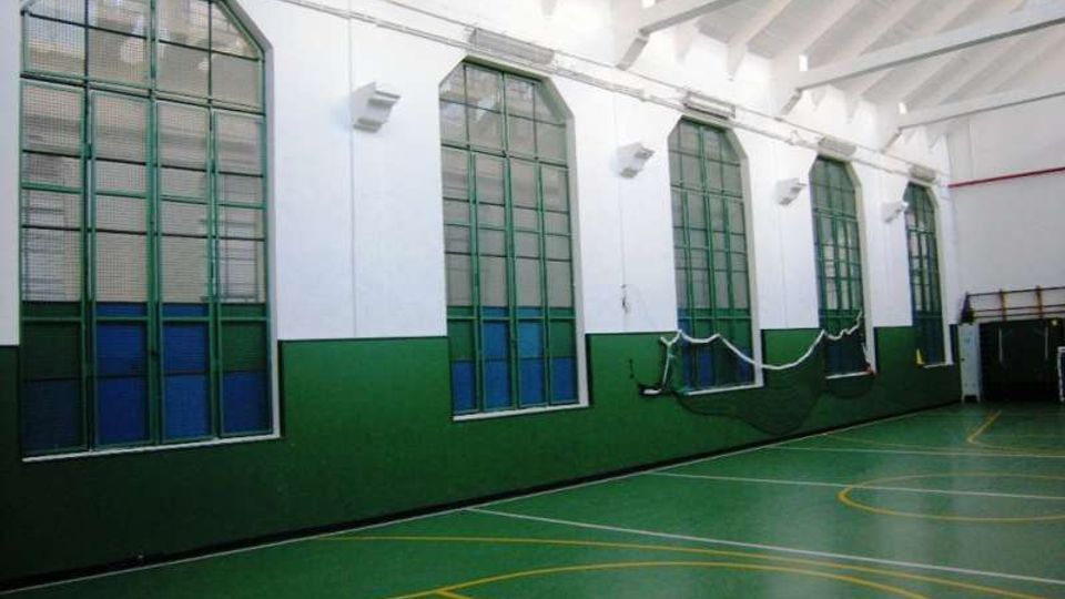 Interno di una palestra con pavimento verde segnato da linee gialle per campi sportivi e pareti bianche con fascia verde. Lungo un lato ci sono alte finestre ad arco con telai verdi e reti appese, sotto un soffitto alto con travi a vista.