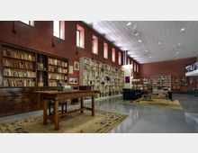 Interno di una biblioteca con pareti in mattoni rossi, alte librerie piene di libri e finestre strette in alto che lasciano entrare luce. In primo piano c’è un grande tavolo in legno su un tappeto, con area lettura sullo sfondo e soffitto alto con illuminazione a pannelli.