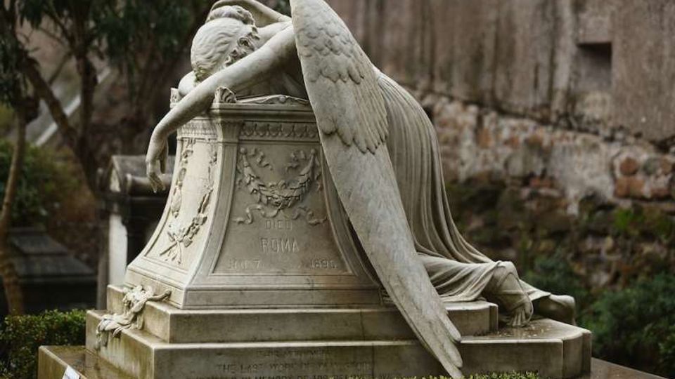 Monumento funerario in marmo in un cimitero, con statua di un angelo sdraiato sul sarcofago e grandi ali scolpite. La base presenta bassorilievi e iscrizioni, circondata da siepi e lapidi sullo sfondo.