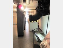 Corridoio interno di un edificio con pavimento piastrellato e soffitto basso, affiancato da colonne scure e illuminazione a parete. Sul lato destro si vedono grandi tubazioni industriali nere e una struttura metallica con pannello informativo.
