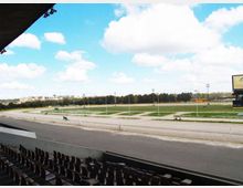 Tribuna coperta con file di sedili affacciata su un ippodromo, con pista ovale e ampi prati interni attraversati da vialetti. Sullo sfondo si vedono alberi, pali dell’illuminazione e un grande tabellone sotto un cielo azzurro con nuvole.