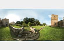Ampio prato all’interno di un complesso storico in pietra, con muretti bassi e una scalinata centrale che conduce a un’area verde con alberi. Ai lati si vedono edifici in muratura e una torre quadrata in pietra, sotto un cielo parzialmente nuvoloso.