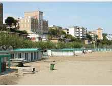 Spiaggia sabbiosa con una fila di cabine e strutture balneari basse dai profili verde acqua, con assi e materiali impilati in primo piano. Sullo sfondo si vede un lungomare con vegetazione e palme e, più in alto, edifici residenziali e un palazzo in stile storico su una collina.