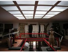 Interno di un atelier o laboratorio di sartoria con grande lucernario a griglia sul soffitto e passerella con ringhiera rossa attorno a un vuoto centrale. Sul fondo sono appesi capi chiari e scuri su una barra, con arredi e attrezzature da lavoro ai lati.