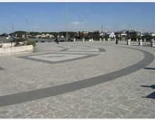 Ampia piazza pavimentata in pietra con motivi geometrici circolari e decorazioni incise, delimitata da lampioni e una balaustra. Sullo sfondo si vedono una strada con parcheggi e edifici bassi sotto un cielo sereno.