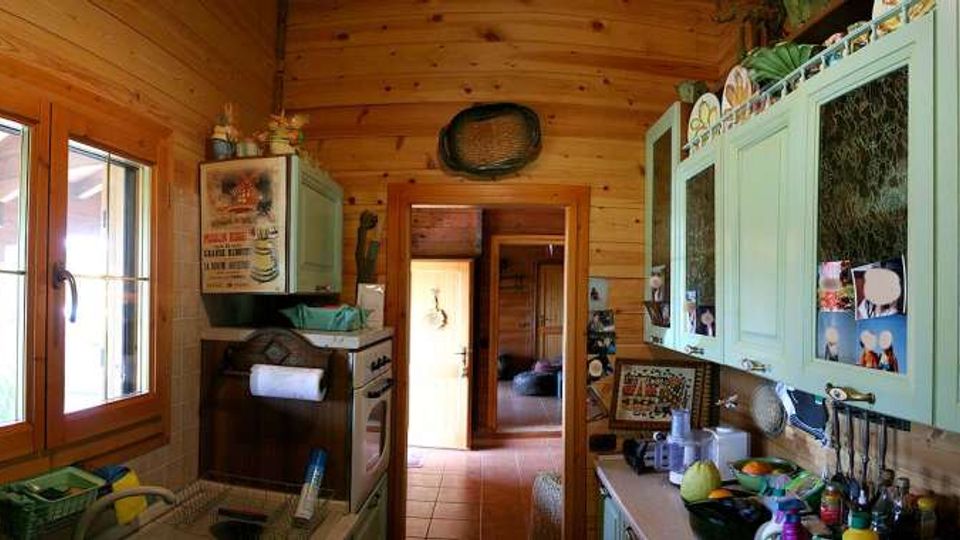 Interno di una cucina rustica in legno con soffitto curvo, finestra a due ante e mobili verde chiaro con ante in vetro. Piano di lavoro con utensili e oggetti, pareti decorate con piastrelle e foto, e porta che conduce a un corridoio con pavimento in piastrelle.