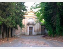 Edificio storico con facciata in muratura e frontone triangolare, con portale centrale incorniciato e decorazioni sopra l’ingresso. Si trova in fondo a un viale sterrato, circondato da alberi e vegetazione fitta ai lati.