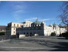 Edificio monumentale in stile classico con facciata chiara, finestre ad arco e grandi cupole verdi, visibile oltre una piazza asfaltata con ringhiere e lampioni. Sullo sfondo si notano colline e un cielo azzurro limpido.