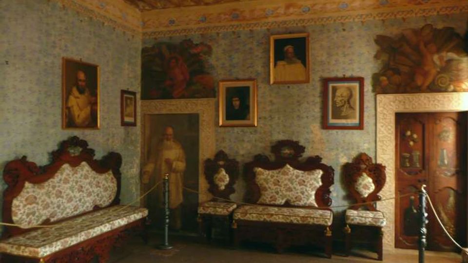 Interno di una sala storica con soffitto a travi di legno decorato, pareti rivestite di carta da parati e numerosi quadri e affreschi. Lungo le pareti sono disposti divani e poltrone imbottite in stile antico, con una vetrina in legno e una corda di delimitazione per i visitatori.