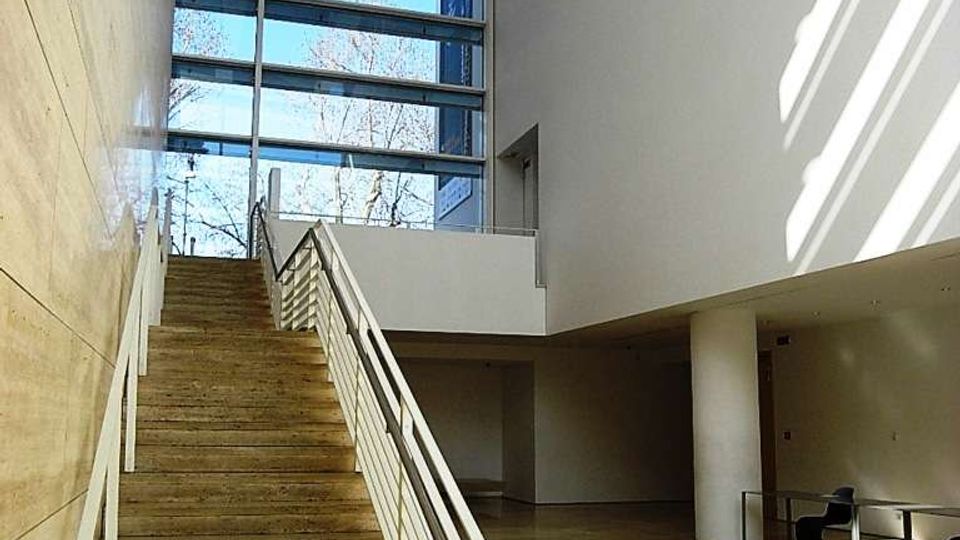 Atrio interno moderno con ampia scala in pietra e corrimano metallici, pavimento in grandi lastre e soffitto alto. Una grande vetrata a tutta altezza illumina lo spazio, con vista su alberi all’esterno.