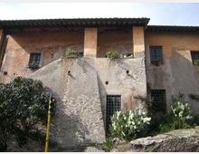 Facciata di un edificio rustico in intonaco e pietra con un grande contrafforte centrale, finestre con inferriate e tetto a falde sporgente. In primo piano vegetazione con cactus e un piccolo semaforo su palo giallo, con muretto di pietra ai bordi.