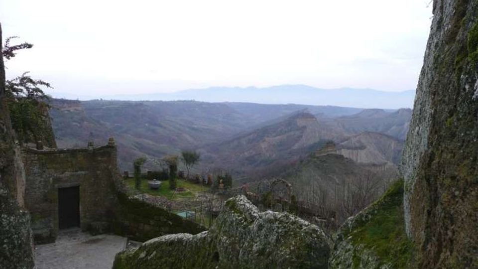 Veduta panoramica di una valle collinare con rilievi e creste in lontananza, osservata da un belvedere in pietra. In primo piano si vedono rocce ricoperte di muschio e un piccolo edificio in muratura con terrazza e muretto.