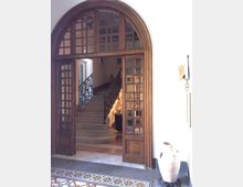 Grande portale interno ad arco in legno con riquadri di vetro, che introduce a un atrio con ampia scala in pietra e ringhiera in ferro battuto. In primo piano si vede un pavimento a mosaico decorato e, a lato, un grande vaso in ceramica contro una parete chiara.