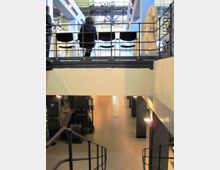 Interno di un museo industriale su due livelli, con soppalco in metallo e ringhiere sopra una grande sala. Al piano terra si vedono macchinari storici e una scala metallica, mentre in alto ci sono sedute e una statua vicino a grandi finestre e travi a vista.