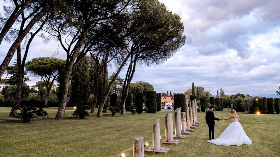 Ampio giardino con prato curato, alti pini a ombrello e siepi potate; una fila di colonne in pietra illuminate segna un percorso verso un edificio in lontananza sotto un cielo nuvoloso.