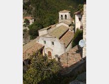 Chiesa in pietra su un pendio, con facciata semplice e tetti in coppi, affiancata da un campanile quadrato con doppie arcate. Sullo sfondo si vede una valle boscosa con alcune case sparse.