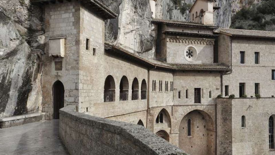 Complesso monastico in pietra addossato a una parete rocciosa, con portico ad archi, finestre strette e tetti in coppi. In primo piano un camminamento con parapetto conduce a un ingresso ad arco e a corpi di fabbrica con piccoli campanili e rosone circolare.