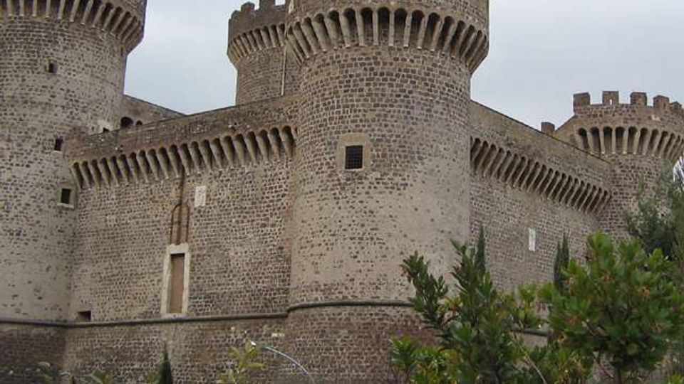 Castello medievale in pietra con mura fortificate e tre grandi torri cilindriche merlate, dotate di beccatelli e piccole feritoie. In primo piano si vede vegetazione arbustiva, con cielo coperto sullo sfondo.
