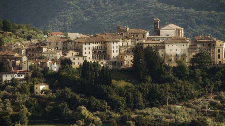 Borgo collinare con case in pietra e intonaco dai tetti in coppi, raccolte attorno a una chiesa con facciata chiara e campanile. In primo piano si vedono campi e uliveti, con boschi e pendii montuosi sullo sfondo.