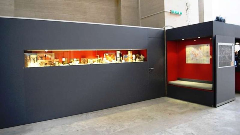 Allestimento museale in uno spazio interno con pareti grigio scuro e una lunga vetrina orizzontale illuminata che espone oggetti. A destra c’è una nicchia con pareti rosse, panca integrata e un pannello espositivo, su pavimento in cemento.