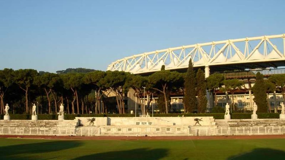 Campo sportivo con pista e prato verde in primo piano, delimitato da una bassa recinzione e da una terrazza con statue in pietra. Sullo sfondo si vedono alberi alti e una grande struttura bianca a traliccio simile a uno stadio sotto un cielo sereno.