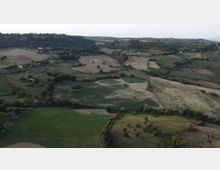 Veduta aerea di una campagna collinare con campi coltivati a parcelle, filari di vigneti e aree verdi intervallate da boschetti. Sentieri e strade sterrate attraversano i terreni, con colline boscate sullo sfondo.