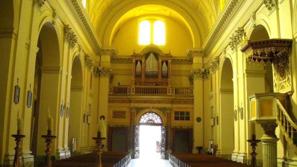 Interno di una chiesa con navata centrale lunga e simmetrica, volta a botte e arcate laterali su colonne. In fondo si vedono la cantoria con grande organo a canne, la porta d’ingresso luminosa e, sulla destra, un pulpito decorato.