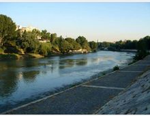 Vista di un fiume che scorre tra sponde alberate, con un’ampia banchina in pietra e una passeggiata lastricata lungo la riva destra. Sullo sfondo si intravedono edifici e vegetazione sotto un cielo sereno.