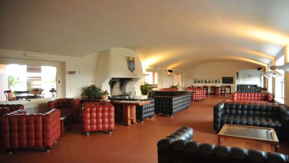 Ampio salone interno con soffitto a volta illuminato, pavimento in piastrelle e grande camino in muratura su una parete. Sono presenti divani Chesterfield in pelle rossa e nera disposti in gruppi con tavolini, con finestre laterali e area bar in fondo alla sala.