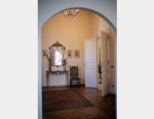 Interno di un ingresso o salotto con grande arco, pavimento in parquet e tappeto orientale; sulla parete un tavolino consolle con specchio decorato e piccoli ritratti incorniciati, con sedia verde accanto. A destra una porta bianca aperta conduce a un’altra stanza, con lampadario a cristalli sospeso in alto.