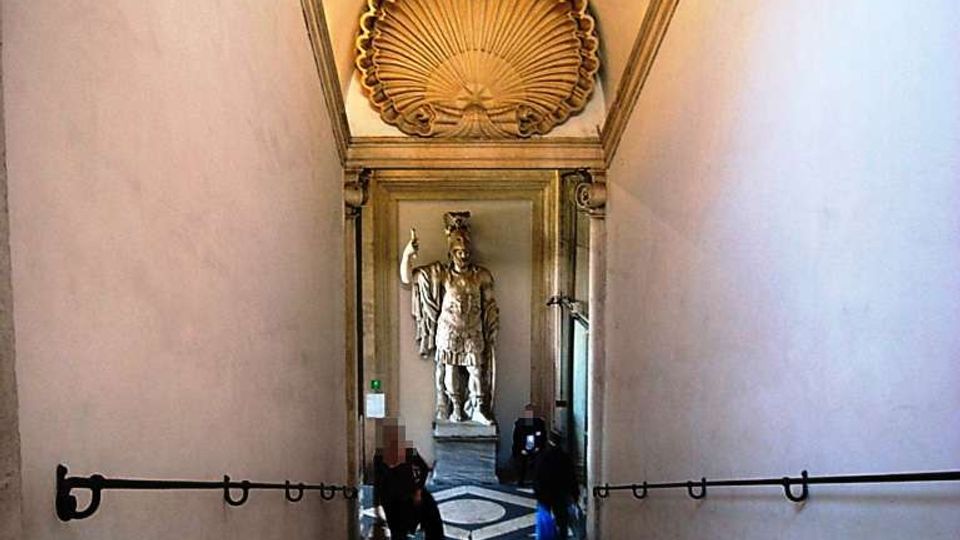 Scalinata in pietra all’interno di un edificio storico, con pareti chiare e corrimano in metallo che conduce a un vestibolo. In fondo si vede una grande statua su piedistallo e un soffitto decorato con una rosetta a conchiglia.