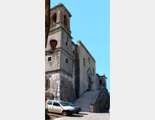 Chiesa in muratura intonacata con campanile a torre e piccole finestre, affacciata su una strada di paese. Una lunga scalinata in pietra conduce all’ingresso laterale, con lampioni e alcune auto parcheggiate in primo piano.