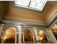 Interno di un edificio storico con cortile o atrio a due livelli, caratterizzato da una serie di arcate e colonne in pietra. In alto si vede un grande lucernario rettangolare con struttura in vetro e cornice decorata che illumina lo spazio.