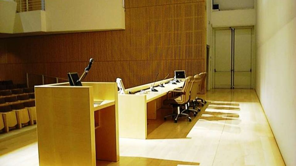 Interno di un’aula magna o sala conferenze con palco in legno, podio con microfono e lungo tavolo con sedie e postazioni. Pareti rivestite in pannelli di legno, balconata laterale e grandi porte sul fondo.