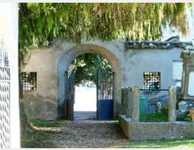 Ingresso con arco in muratura in un muro intonacato, con cancello metallico aperto che conduce a un’area esterna alberata. Ai lati dell’arco si vedono piccole finestre con grata e un vialetto di ghiaia sotto rami pendenti.